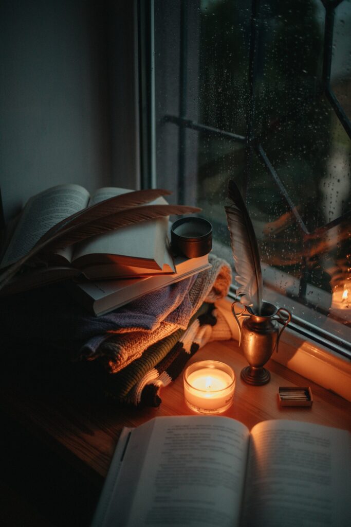 A candle and a pile of books on a window sill on a dark, rainy evening